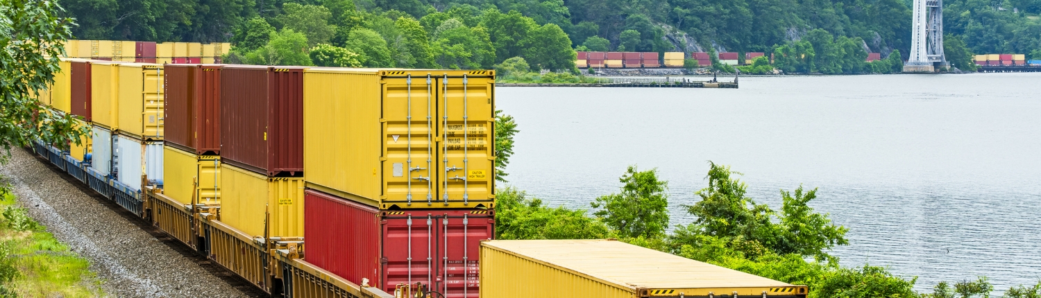 Container stack freight train rounding curve along Hudson River edge with Bear Mountain bridge